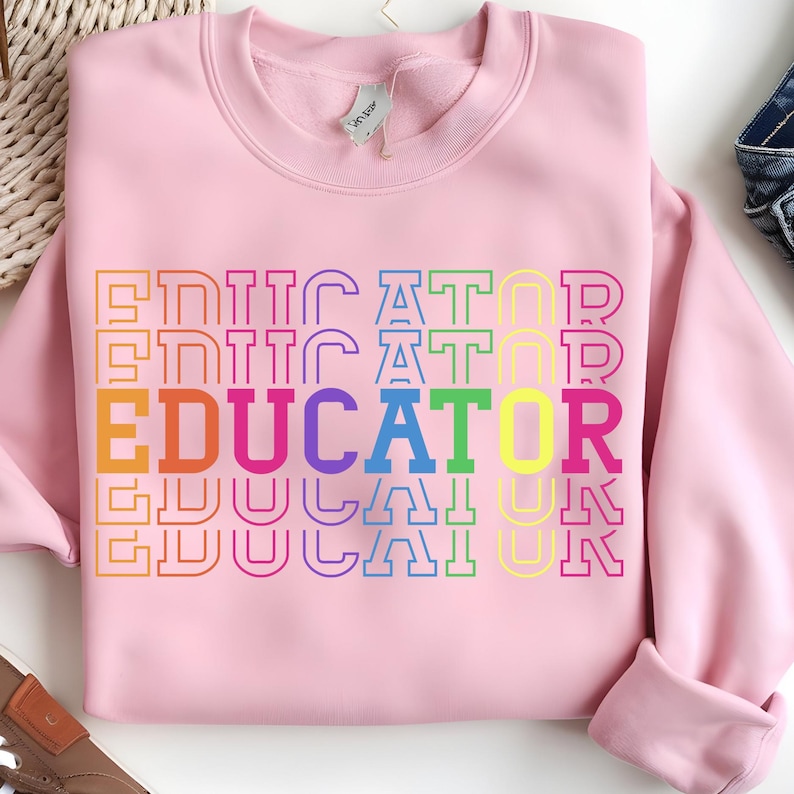 May include: A light pink sweatshirt with the word "EDUCATOR" printed in a rainbow of colors. The word is stacked vertically and outlined in various hues, creating a vibrant and eye-catching design. The sweatshirt has a crew neck and long sleeves.