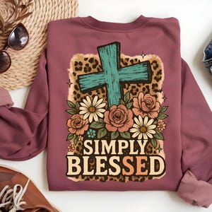 May include: Mauve sweatshirt with a graphic design featuring a turquoise cross, flowers, and the words "SIMPLY BLESSED" in a decorative font. The design includes a leopard print background.