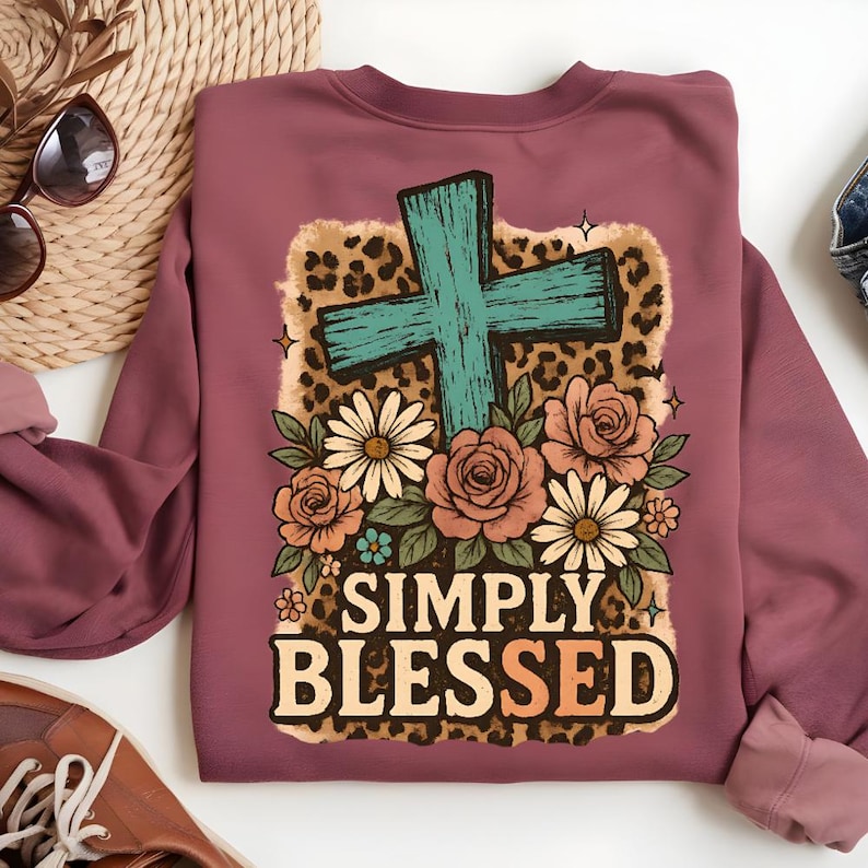 May include: Mauve sweatshirt with a graphic design featuring a turquoise cross, flowers, and the words "SIMPLY BLESSED" in a decorative font. The design includes a leopard print background.