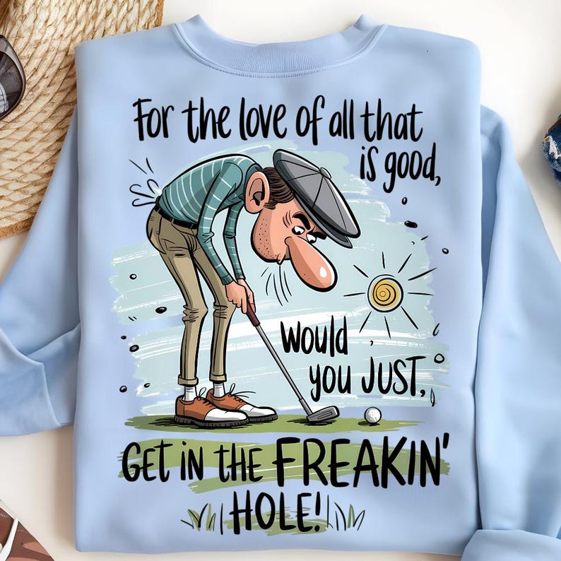 Puede incluir: Sudadera azul claro con un gr&aacute;fico humor&iacute;stico con tem&aacute;tica de golf. El dibujo animado representa a un golfista con el texto: "For the love of all that is good, would you JUST, GET IN THE FREAKIN' HOLE!" Tambi&eacute;n hay una pelota de golf y un sol.