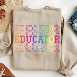 May include: A beige sweatshirt with the word "EDUCATOR" in a rainbow of colors. The word is stacked vertically and repeated multiple times. The sweatshirt has a crew neck and long sleeves.