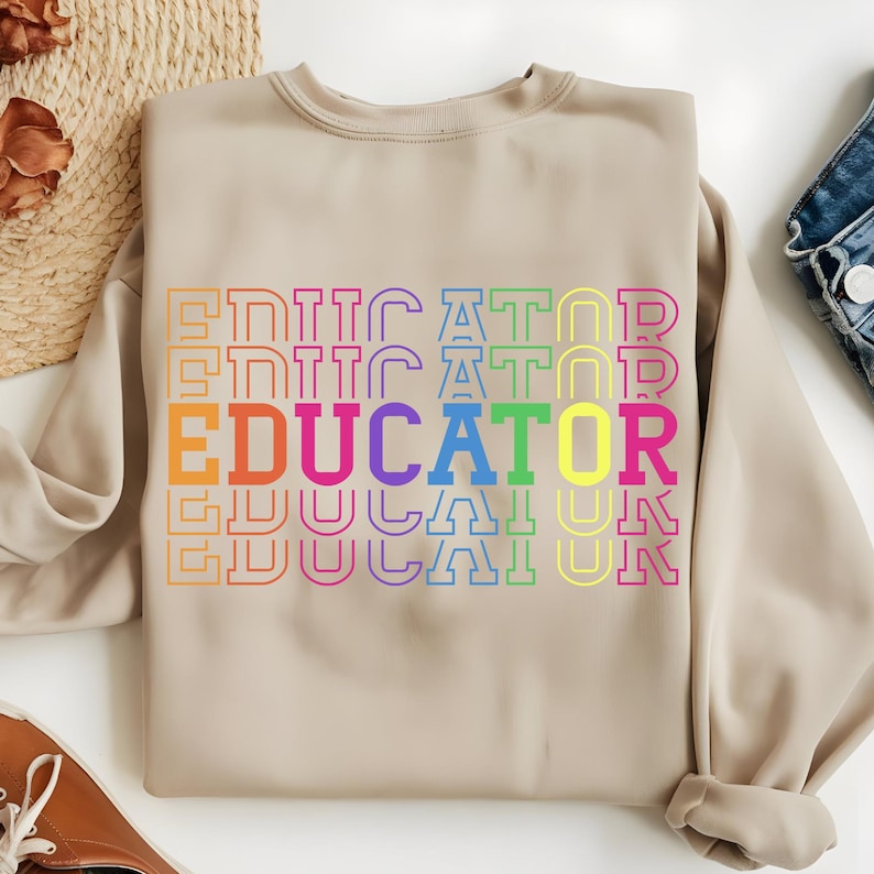 May include: A beige sweatshirt with the word "EDUCATOR" in a rainbow of colors. The word is stacked vertically and repeated multiple times. The sweatshirt has a crew neck and long sleeves.