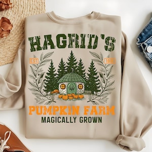 May include: Beige sweatshirt with a green and orange graphic. The graphic features the text "Hagrid's Pumpkin Farm" with an illustration of a small house, pumpkins, and trees. The text "Magically Grown" is also included.