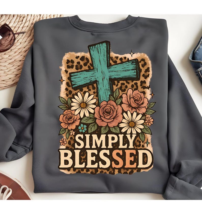 May include: A gray sweatshirt featuring a teal cross, floral arrangement, and leopard print background. The words "SIMPLY BLESSED" are printed in a bold, orange font. The design is centered on the front of the sweatshirt.
