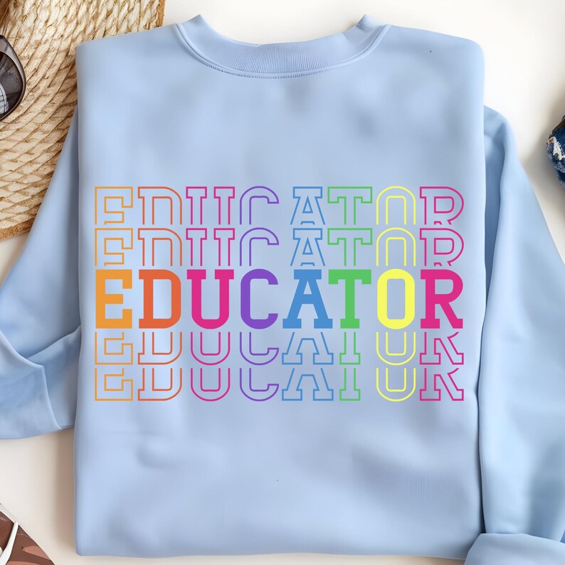 May include: Light blue sweatshirt with the word "EDUCATOR" in large, colorful letters. The word is stacked vertically with a rainbow gradient, and the word "EDUCATOR" is in a bold, bright orange color.