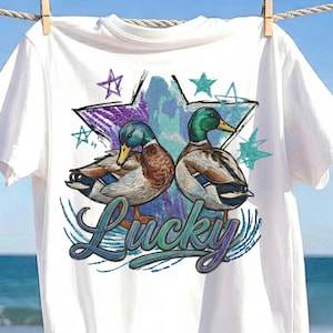 Puede incluir: Una camiseta blanca de manga corta con un gráfico de dos patos reales mirando en direcciones opuestas. Detrás de los patos hay una estrella grande con texturas moradas y azules. La palabra Lucky está impresa en cursiva bajo los patos.