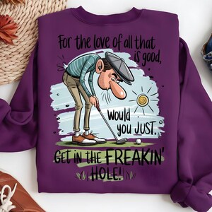 Puede incluir: Una sudadera morada con un dise&ntilde;o de golf. La ilustraci&oacute;n muestra a un golfista con el texto: "For the love of all that is good, Would you JUST, GET IN THE FREAKIN' HOLE!" Incluye una pelota de golf y un sol.