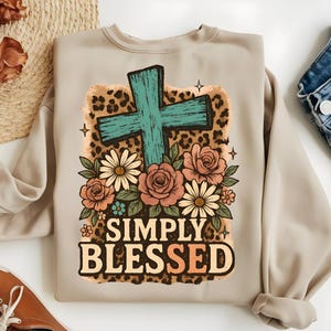 May include: Beige sweatshirt featuring a turquoise cross, surrounded by flowers and a leopard print background. The words "SIMPLY BLESSED" are printed in a bold, stylized font. The design includes daisies, roses, and small floral accents.