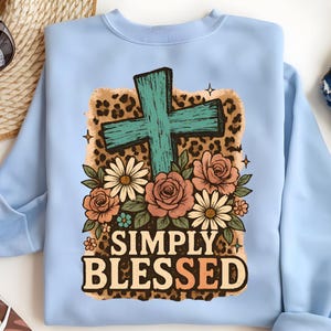 May include: Light blue sweatshirt featuring a teal cross, surrounded by roses and daisies. The design includes a leopard print background and the words "SIMPLY BLESSED" in a decorative font. A casual, faith-based apparel item.