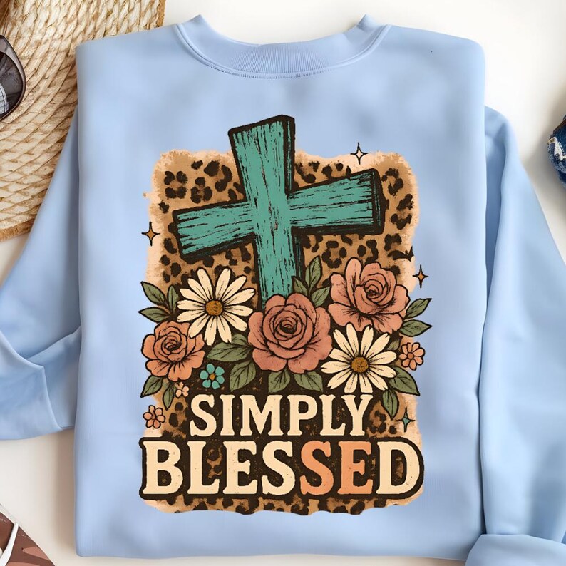 May include: Light blue sweatshirt featuring a teal cross, surrounded by roses and daisies. The design includes a leopard print background and the words "SIMPLY BLESSED" in a decorative font. A casual, faith-based apparel item.