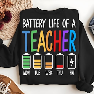 Pode incluir: Camisola preta com o texto "BATTERY LIFE OF A TEACHER" em branco e letras coloridas. Abaixo, ícones de bateria mostrando a vida útil da bateria de segunda a sexta-feira. Gola redonda.