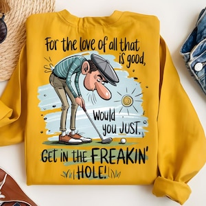 Puede incluir: Sudadera amarilla mostaza con un gr&aacute;fico humor&iacute;stico con tem&aacute;tica de golf. La ilustraci&oacute;n representa a un golfista con una nariz larga, inclinado sobre un palo de golf, con el texto "For the love of all that is good, would you JUST, GET IN THE FREAKIN' HOLE!"