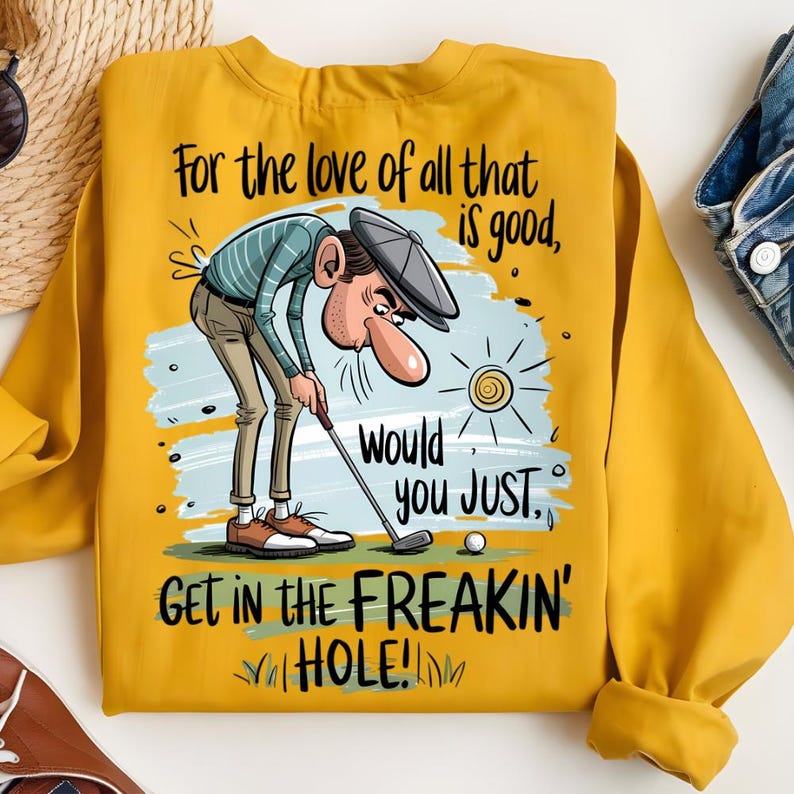 Puede incluir: Sudadera amarilla mostaza con un gr&aacute;fico humor&iacute;stico con tem&aacute;tica de golf. La ilustraci&oacute;n representa a un golfista con una nariz larga, inclinado sobre un palo de golf, con el texto "For the love of all that is good, would you JUST, GET IN THE FREAKIN' HOLE!"