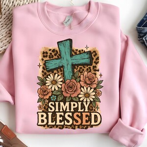 May include: A light pink sweatshirt featuring a teal cross design with floral accents and the words "SIMPLY BLESSED" in a decorative font. The cross is set against a leopard print background, with roses and daisies.