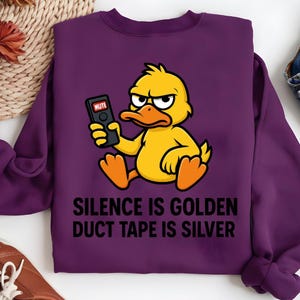 Könnte beinhalten: Ein lila Sweatshirt mit einer Cartoon-Ente, die eine Fernbedienung mit dem Wort "MUTE" hält. Der Text "SILENCE IS GOLDEN DUCT TAPE IS SILVER" ist darunter gedruckt. Die Ente ist gelb mit orangefarbenen Füßen und schwarzer Umrandung.