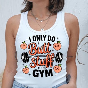 Op de afbeelding: Witte tanktop met de tekst "I Only Do Butt Stuff At The Gym". Het ontwerp bevat een halter, perziken en sterren. De tekst en afbeeldingen zijn in zwart en oranje.