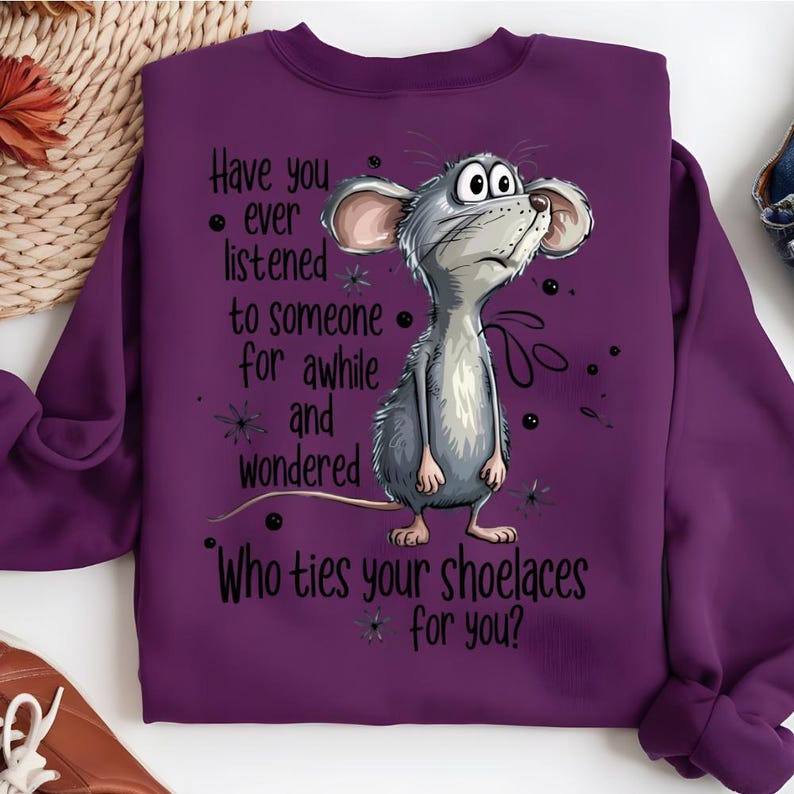 May include: A purple sweatshirt with a cartoon illustration of a gray mouse. The mouse is looking up with a curious expression. Text on the shirt reads "Have you ever listened to someone for awhile and wondered Who ties your shoelaces for you?"