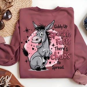 Pode incluir: Um moletom bord&ocirc; com um desenho de burro de desenho animado e o texto "Giddy Up Sparkle Farts There's Chaos to Spread." O burro &eacute; cinza com crina e cauda pretas, em um fundo de nuvem rosa com estrelas e cora&ccedil;&otilde;es.