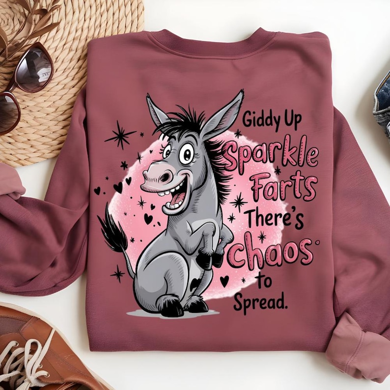Pode incluir: Um moletom bord&ocirc; com um desenho de burro de desenho animado e o texto "Giddy Up Sparkle Farts There's Chaos to Spread." O burro &eacute; cinza com crina e cauda pretas, em um fundo de nuvem rosa com estrelas e cora&ccedil;&otilde;es.