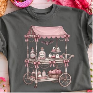 May include: A charcoal grey t-shirt featuring a graphic of a pink cart with a canopy and bows. The cart displays glass bottles, ghost-shaped treats, macarons, and a basket of pumpkins. A bat is illustrated above the cart, and the bottles have text.