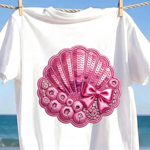 May include: A white short-sleeved t-shirt featuring a large, circular pink seashell graphic on the front. The design is embellished with shiny pink sequins, textured circular accents, small clear rhinestones, and a prominent pink satin bow.