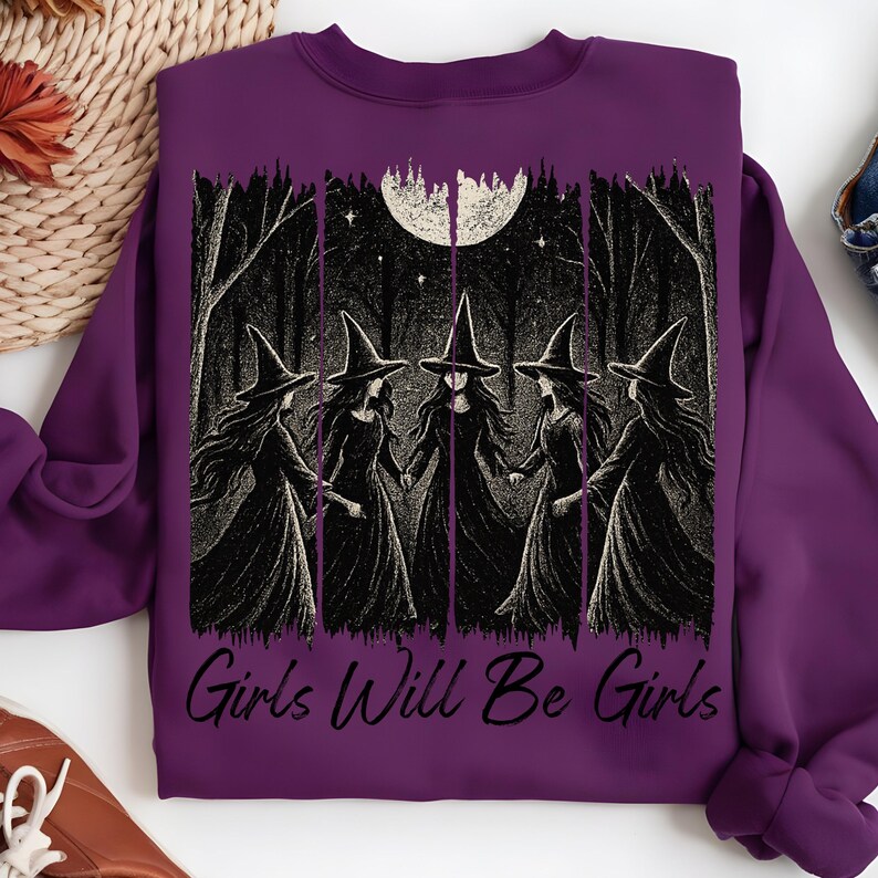 May include: A purple sweatshirt with a graphic of five witches in a forest under a full moon. The phrase "Girls Will Be Girls" is printed below the image. The witches are wearing black dresses and hats. The sweatshirt is made of a soft material.