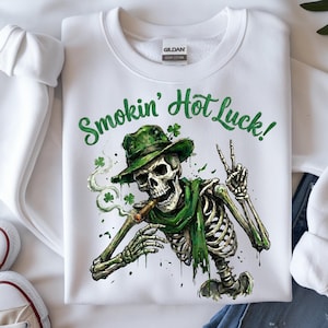 Op de afbeelding: Witte sweatshirt met een afbeelding van een skelet met een groene hoed en sjaal, een sigaar rokend. De tekst "Smokin' Hot Luck!" staat in het groen boven het skelet. Het skelet maakt het vredesteken.