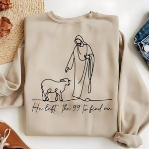 May include: Beige sweatshirt with a black line drawing of a figure with a robe and a lamb. The text "He left the 99 to find me" is printed in black at the bottom of the sweatshirt. The sweatshirt is made of a soft material.