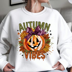 May include: A white crewneck sweatshirt featuring a graphic of an orange jack-o'-lantern with a purple bow on top. The design includes brown and orange maple leaves, black stars, and the text "AUTUMN VIBES" in green and orange dripping letters.