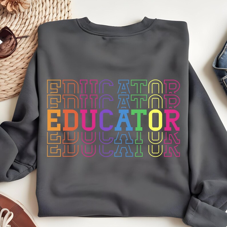 May include: Dark gray sweatshirt with the word "EDUCATOR" in a rainbow of colors. The word is stacked vertically multiple times. The sweatshirt is a crew neck style.