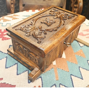 Könnte beinhalten: Eine rechteckige Holzschatulle mit dunkelbraunem Finish, die auf dem Deckel und der Vorderseite mit detaillierten floralen Schnitzereien verziert ist. Die Schatulle hat einen kleinen Metallschlüssel an der Vorderseite und einen erhöhten Sockel.