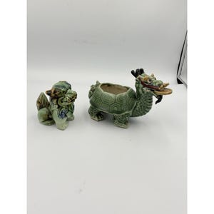 May include: Two ceramic figurines: a green Foo Dog and a dragon-turtle planter. The Foo Dog is light green with detailed features. The dragon-turtle planter has a textured shell and a dragon head, holding a coin in its mouth.