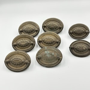 May include: A collection of eight antique brass drawer pulls. Each oval pull features an ornate, embossed design with a central eye-shaped detail and a decorative border. The pulls have a weathered, aged appearance.
