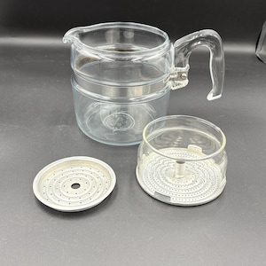 May include: A clear glass coffee percolator with a handle, a lid, and a filter basket. The percolator is made of clear glass and has a silver metal band around the middle. The filter basket and lid are also visible.