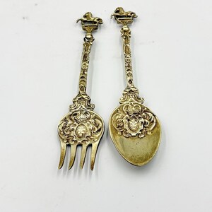 May include: Two ornate, antique silver-colored serving utensils. One is a fork with three tines, and the other is a spoon. Both have detailed floral and crown designs, with a horse figure at the top of the handles. The utensils are likely for serving.