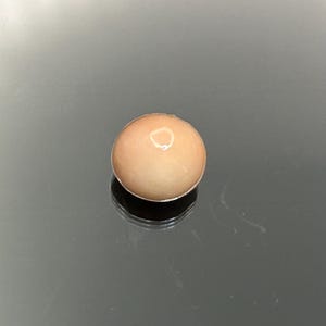 May include: A single, smooth, round, peach-colored cabochon gemstone with a polished, reflective surface. The stone features a soft, warm hue and a domed top, displaying a subtle light reflection on its curved, opaque, and uniform exterior.