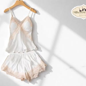 Könnte beinhalten: Ein weißes Seiden-Pyjama-Set mit pfirsichfarbener Spitze, bestehend aus einem Camisole-Top und Shorts, hängt an einem Holzbügel. Das Set wird vor einem weißen Hintergrund mit Schatten präsentiert. Das Logo "SilkenSecrets" ist sichtbar.