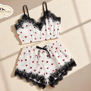 May include: White satin lingerie set with red heart patterns, black lace trim, and a black bow. The set includes a camisole top with thin straps and shorts. The logo "SilkenSecrets by Lior" is visible in the upper left corner.