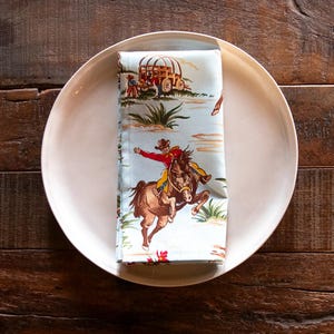 May include: A folded light blue napkin with a Western-themed print featuring cowboys, horses, and a covered wagon. The napkin is placed on a white plate, set against a dark wood background. The design includes red, brown, and yellow accents.