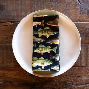 May include: A folded black napkin with a colorful fish pattern rests on a white plate. The fish are various species, including perch and trout, with orange, yellow, and green hues. The plate sits on a dark wooden surface.