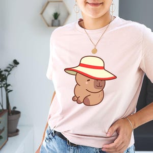 May include: Light pink t-shirt featuring a cartoon capybara wearing a cream-colored hat with a red band. The capybara is brown and has a cute, friendly expression. The shirt is made of soft material and is suitable for casual wear.