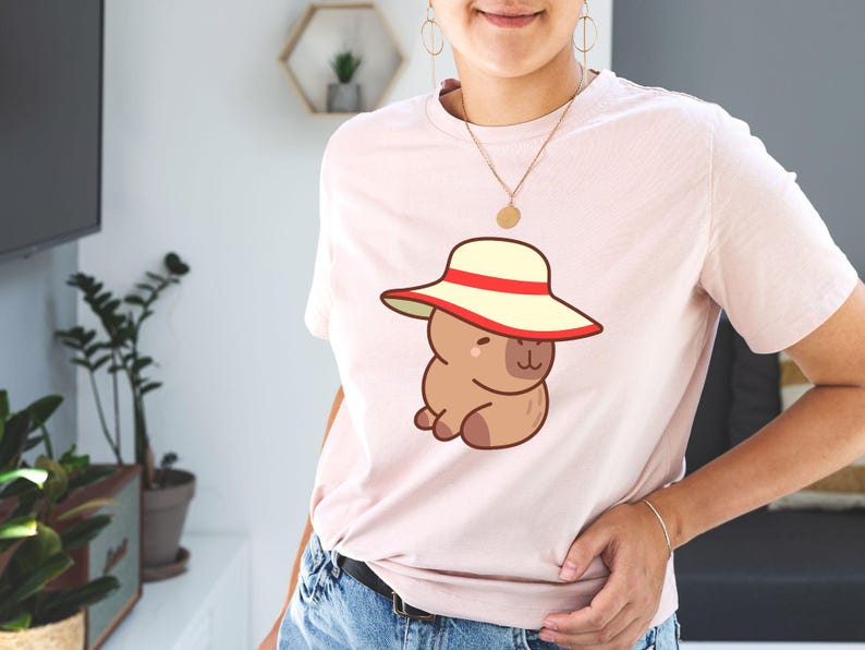 May include: Light pink t-shirt featuring a cartoon capybara wearing a cream-colored hat with a red band. The capybara is brown and has a cute, friendly expression. The shirt is made of soft material and is suitable for casual wear.