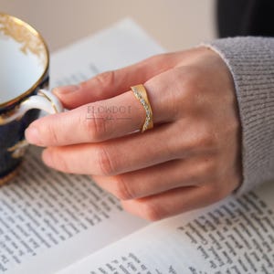May include: A gold ring with a curved band and a row of small clear stones. The ring is on a finger.