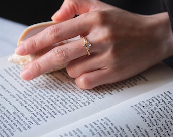 May include: A gold ring with a small, light-colored gemstone is worn on a hand. The ring has a delicate, twisted band. The hand is holding a seashell and resting on an open book. The word "FLOWDOT" is visible on the image.