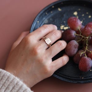 May include: A gold ring featuring an ornate, rectangular face with intricate engraved detailing and a scalloped edge. The band is slender and polished, showcasing a metallic finish that catches the light, worn on a finger near a cluster of red grapes.