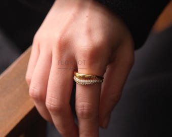 May include: Two gold rings on a finger. The top ring is a simple band. The bottom ring is a curved band with small clear stones.