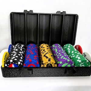 May include: A black plastic carrying case containing five stacks of circular poker chips in black, blue, yellow, purple, and green. Each stack features a distinct graphic design on the face, with additional loose chips placed at the ends.