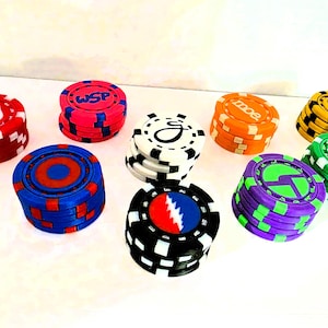 May include: A collection of eight stacks of circular poker chips in various colors including red, blue, pink, white, orange, yellow, green, black, and purple. Each stack features unique graphic designs, logos, or symbols on the top surface.