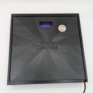 May include: Black square-shaped base with a textured surface and a small purple rectangle in the center. The base has the text "Starzy Creations" printed on it. A silver coin is sitting on the base.