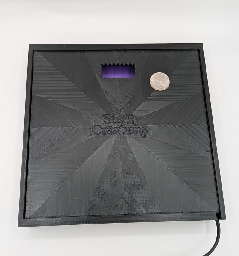 May include: Black square-shaped base with a textured surface and a small purple rectangle in the center. The base has the text "Starzy Creations" printed on it. A silver coin is sitting on the base.